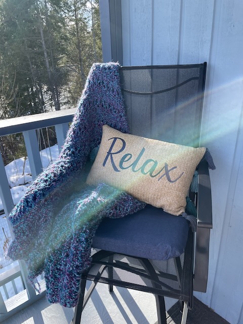 An outdoor chair with a beautiful knit blanket and a tan throw pillow that says "relax"